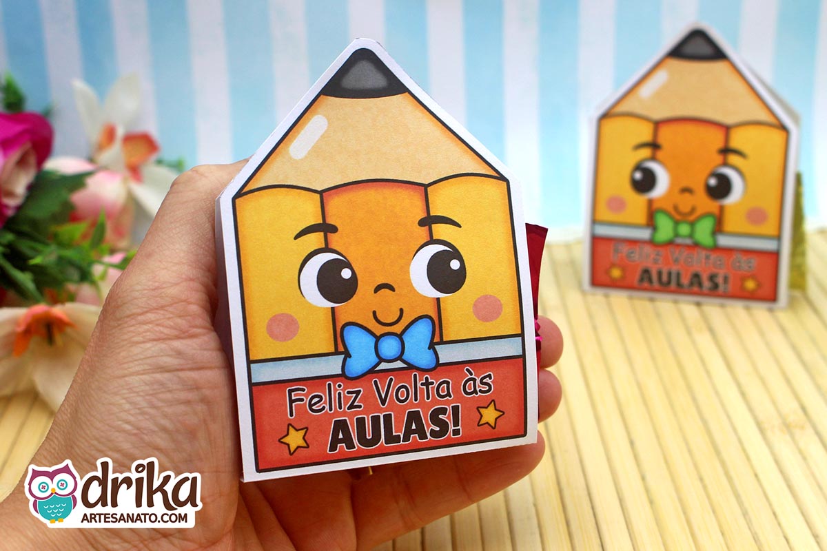Mão segurando um porta-bombom de lápis amarelo com rosto fofo, gravatinha azul e a frase "Feliz Volta às Aulas"