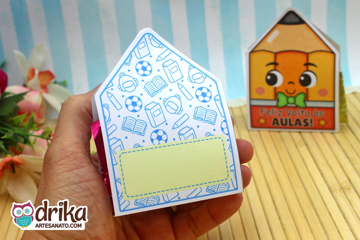 Mão segurando o verso de um porta-bombom lápis com estampa de material escolar e espaço pontilhado para mensagem