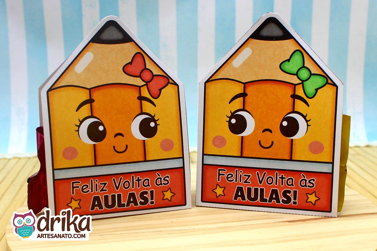 Dois porta-bombons de papel em formato de lápis com rostinhos fofos. Um tem laço vermelho e o outro laço verde, ambos com a frase "Feliz Volta às Aulas"