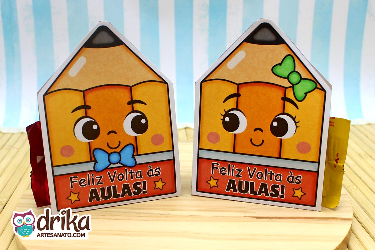 Dois porta-bombons de papel em formato de lápis com rostinhos fofos. Um tem gravatinha azul e o outro laço verde, ambos com a frase "Feliz Volta às Aulas"