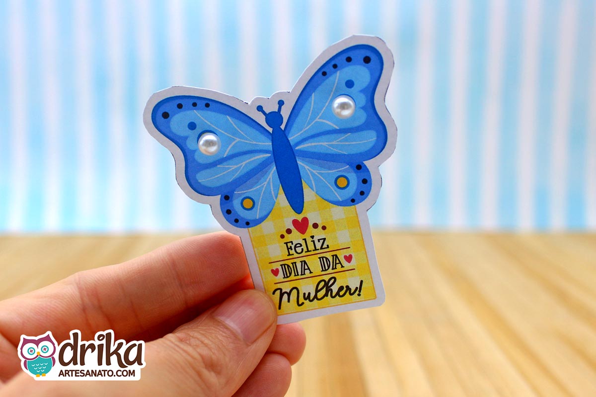 Mão segurando cartão de Dia da Mulher em formato de borboleta azul com duas pérolas brancas nas asas e fundo xadrez amarelo