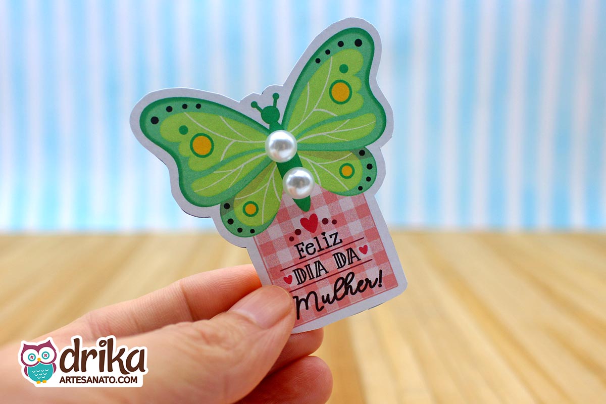 Mão segurando cartão de Dia da Mulher em formato de borboleta verde com duas pérolas brancas nas asas e fundo xadrez rosa