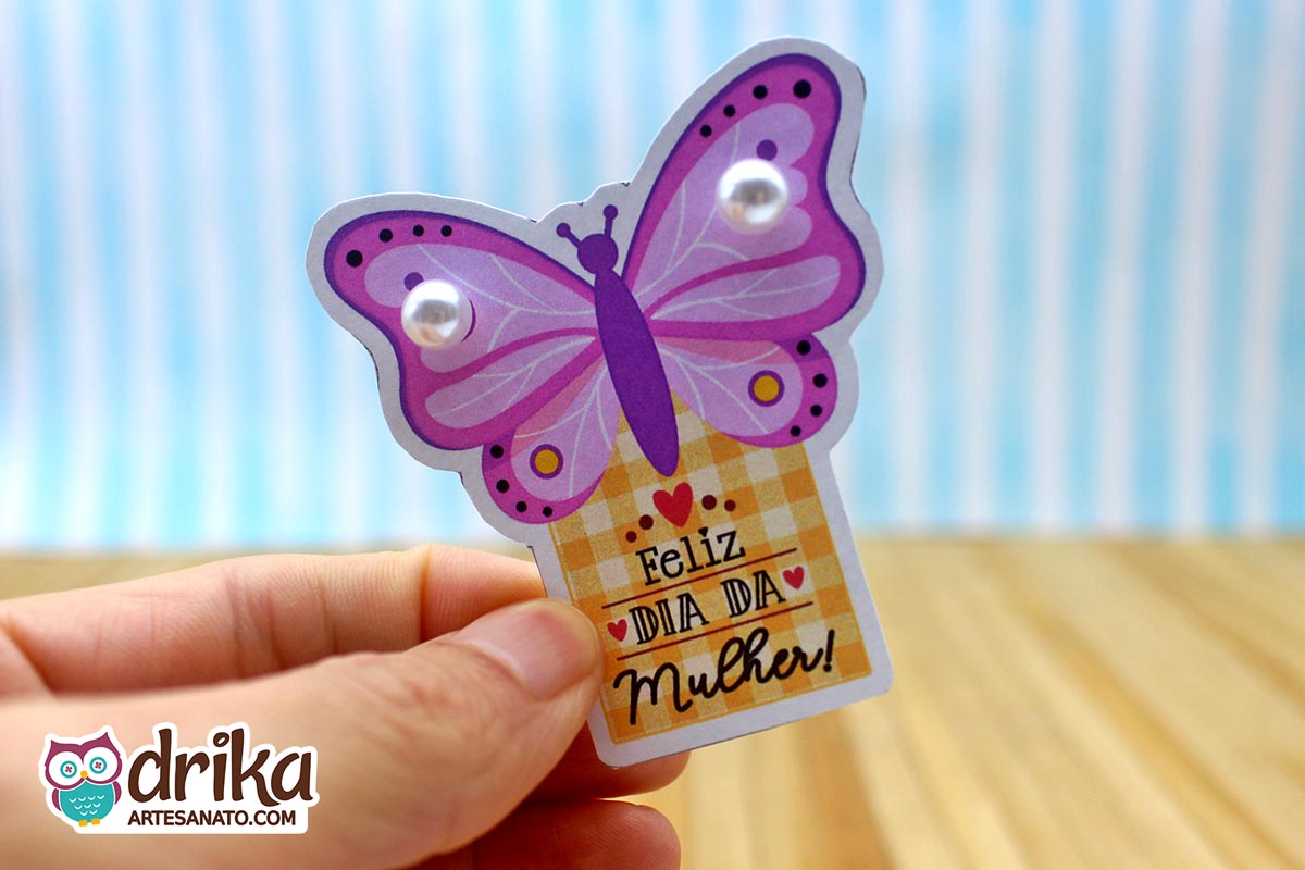 Mão segurando cartão de Dia da Mulher em formato de borboleta lilás com duas pérolas brancas nas asas e fundo xadrez amarelo