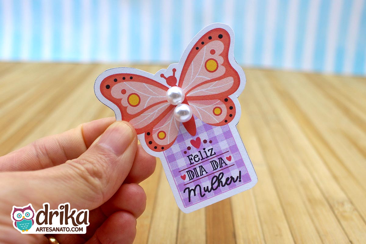 Mão segurando um cartão de borboleta rosa coral com duas pérolas brancas nas asas e a frase "Feliz Dia da Mulher" em fundo xadrez lilás