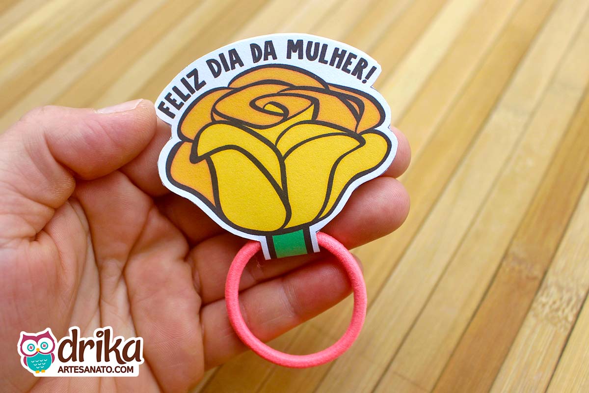 Mão segurando uma lembrancinha de rosa porta xuxinha de papel amarelo com lacinho laranja