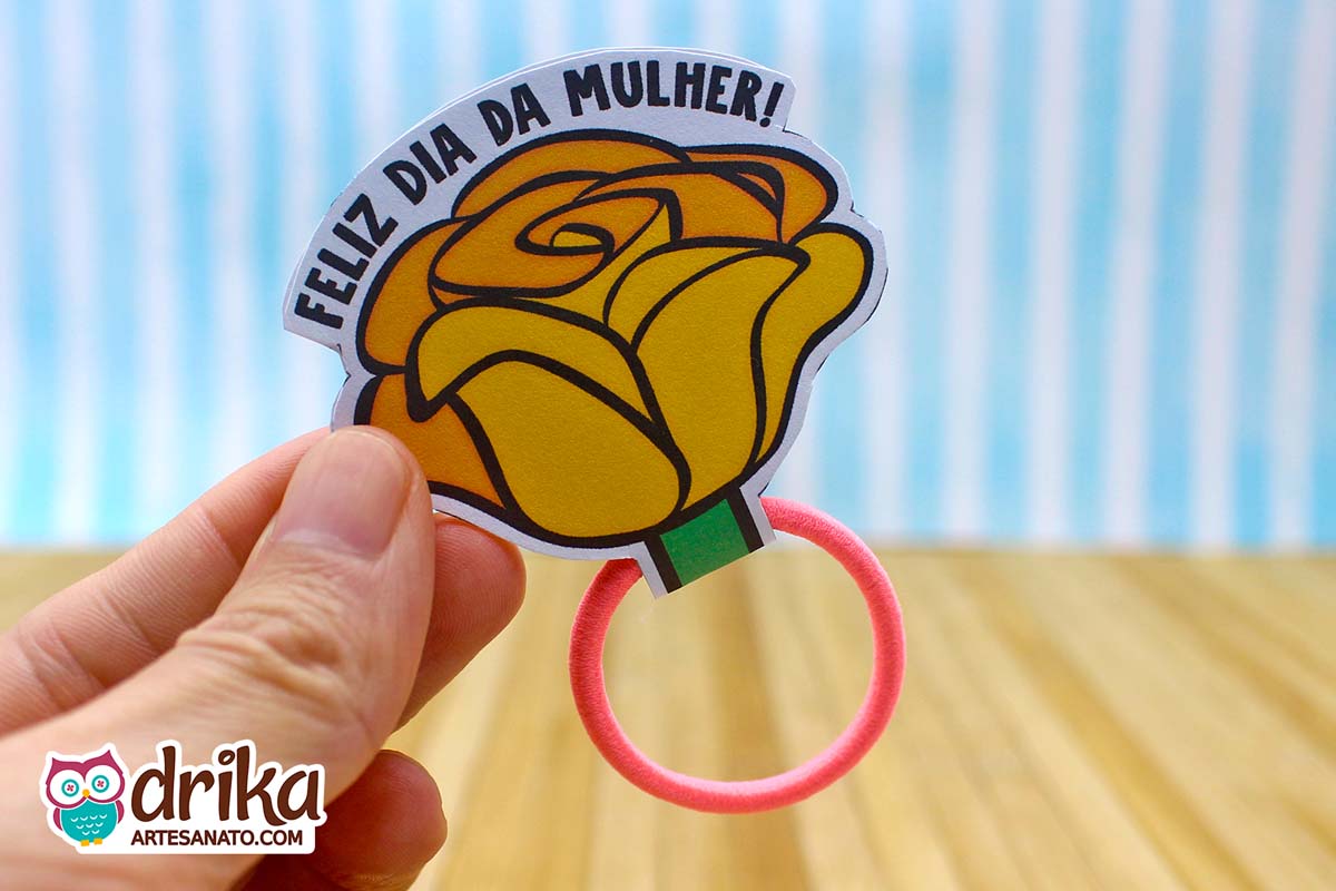 Mão segurando lembrancinha de Dia da Mulher em formato de rosa amarela de papel com frase e xuxinha rosa claro