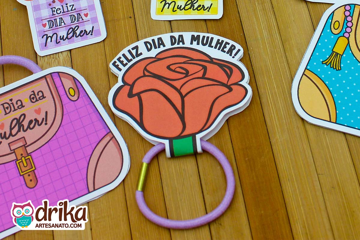 Rosa porta xuxinha laranja de papel com frase "Feliz Dia da Mulher" e xuxinha lilás, entre outras lembrancinhas