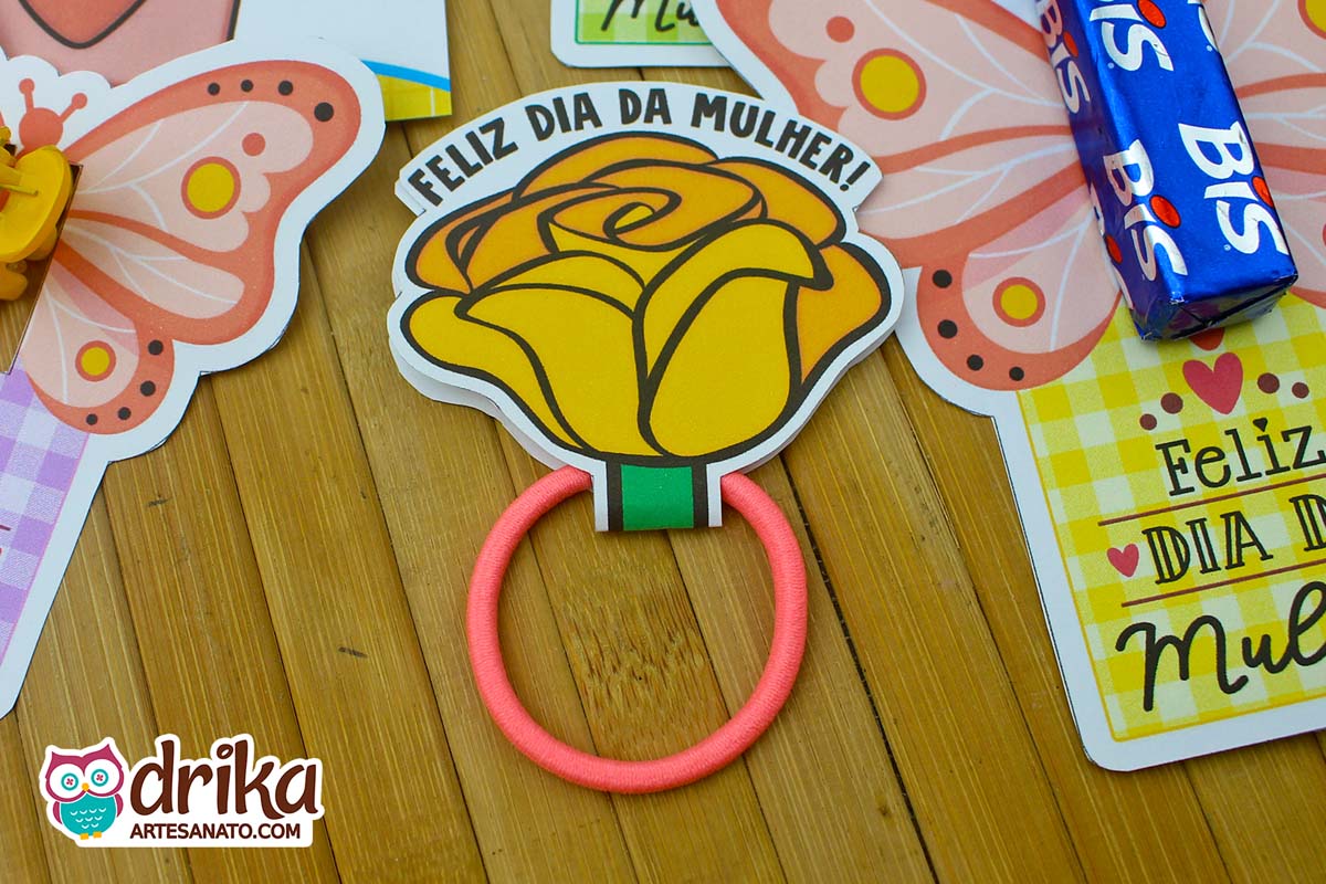 Rosa porta xuxinha amarela de papel com frase "Feliz Dia da Mulher" e xuxinha rosa, cercada por outras lembrancinhas