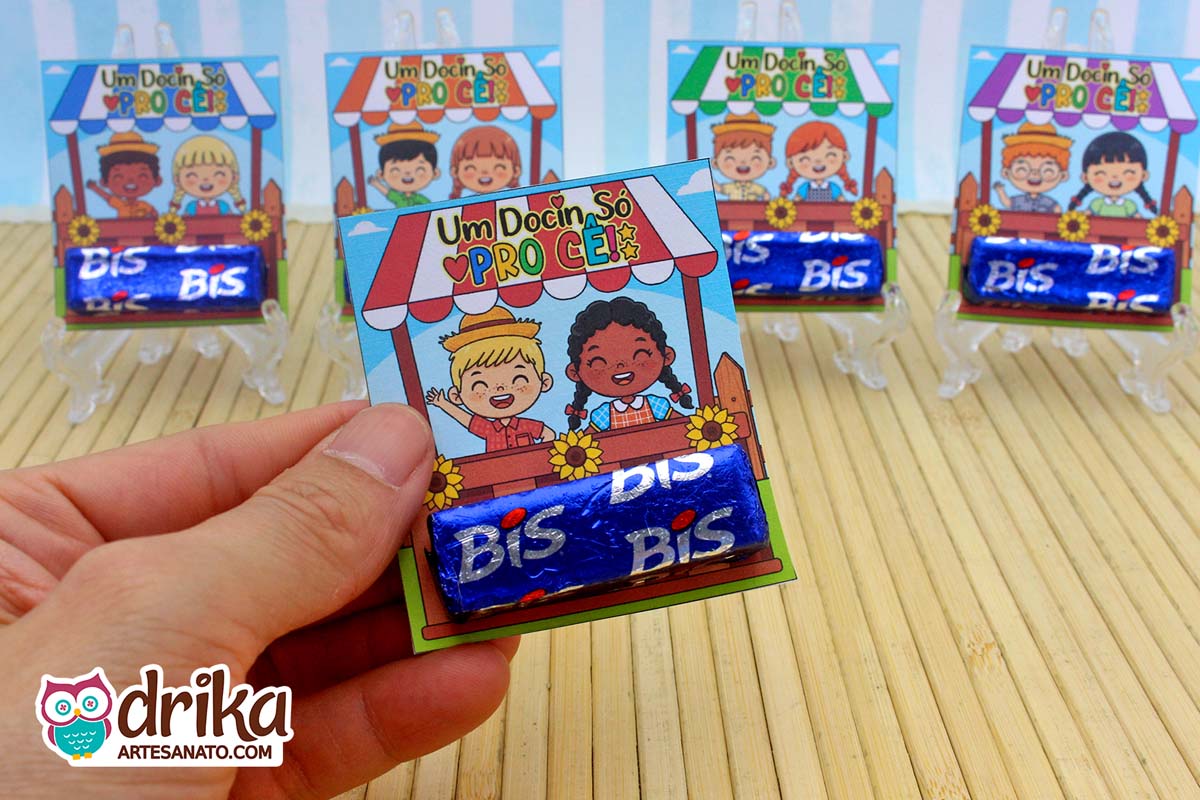 Mão segurando porta-bis caipira com menino loiro e menina negra em barraquinha, girassóis e dois chocolates Bis azuis encaixados