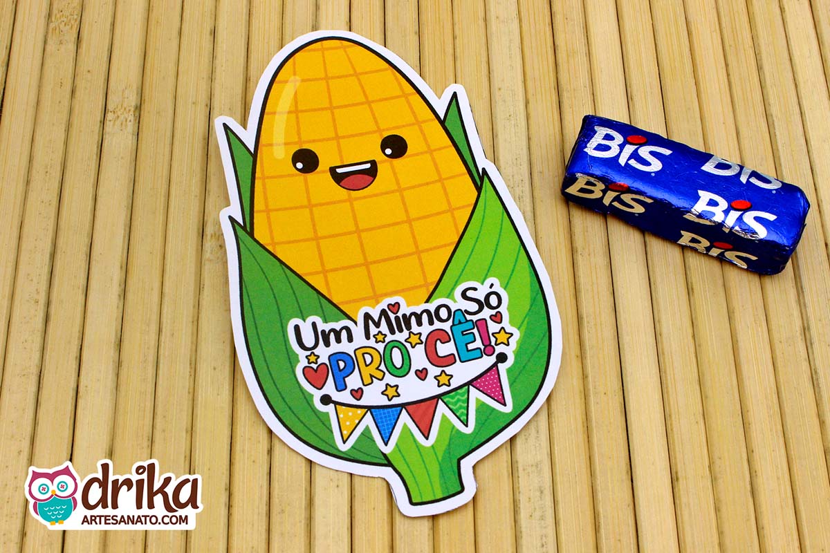 Porta-bis de milho kawaii de papel, amarelo e verde, com frase "Um Mimo Só Pro Cê!" e bandeirinhas, ao lado de um chocolate Bis azul