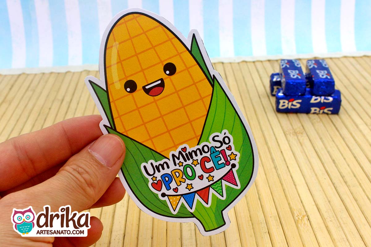 Mão segurando porta-bis de milho kawaii de papel, amarelo e verde, com frase festiva, sobre fundo de bambu e Bis desfocados