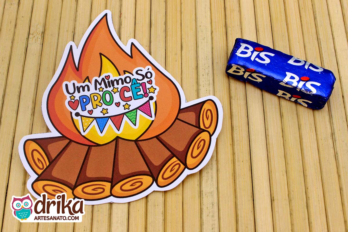 Porta-bis de papel com desenho de fogueira junina e um chocolate Bis azul ao lado sobre superfície de bambu