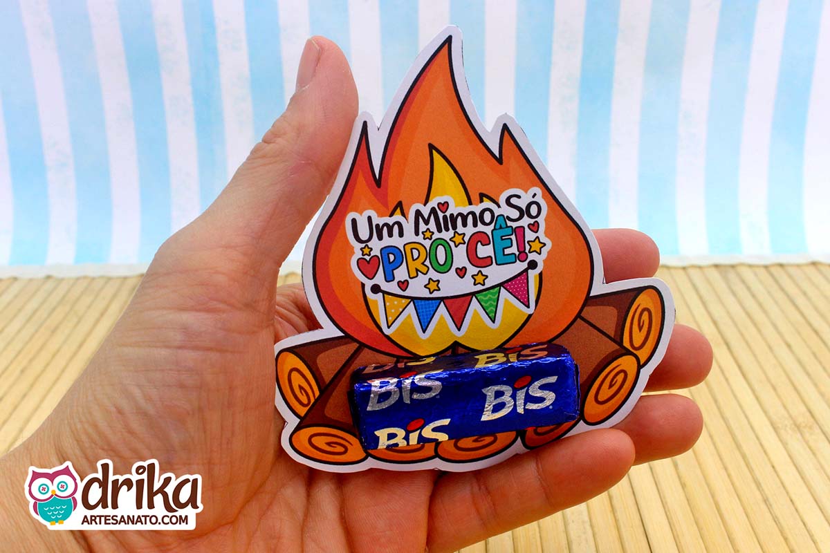 Mão segurando porta-bis de papel com desenho de fogueira e um chocolate Bis azul encaixado nas toras