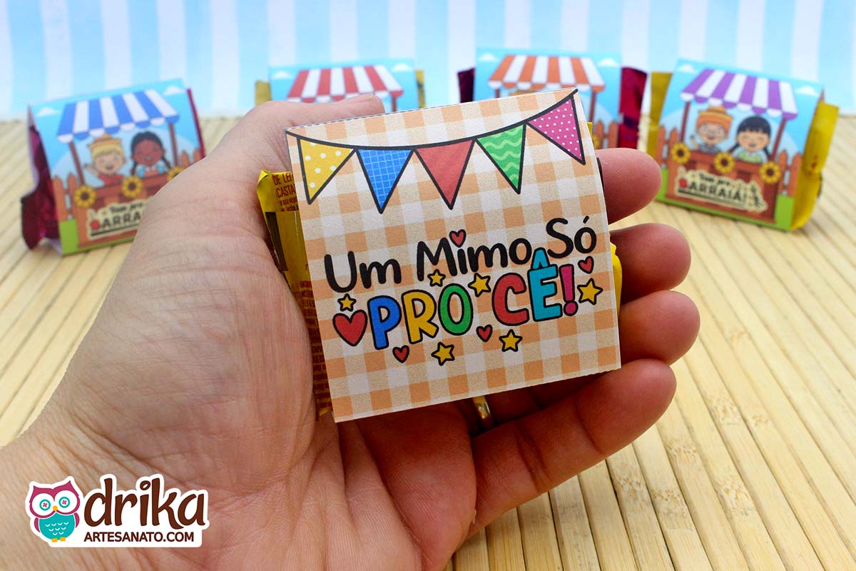 Mão segurando o verso de porta-bombom de papel com fundo xadrez, bandeirinhas juninas e a frase "Um Mimo Só Pro Cê!"