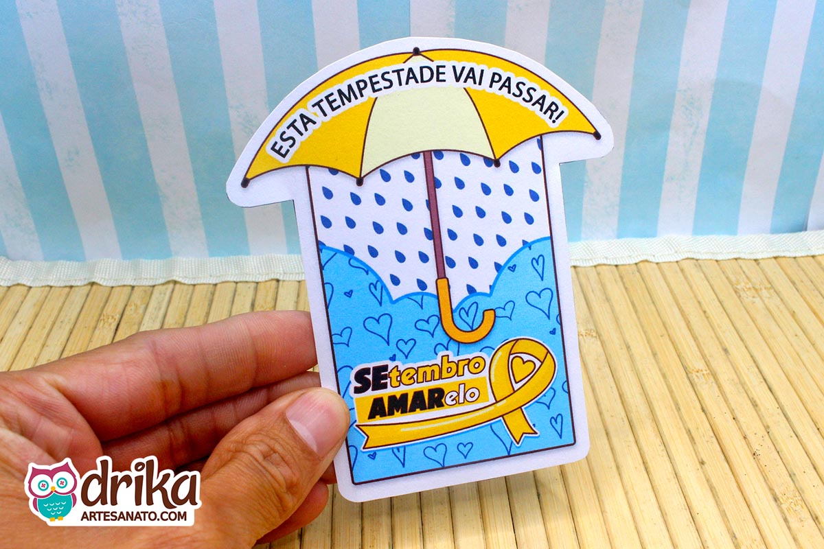 Mão segurando cartão de papel em formato de guarda-chuva com estampa de corações azuis e laço amarelo