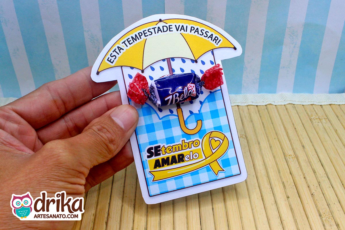 Mão segurando cartão de papel com desenho de guarda-chuva amarelo e gotas de chuva, com uma bala 7Belo fixada