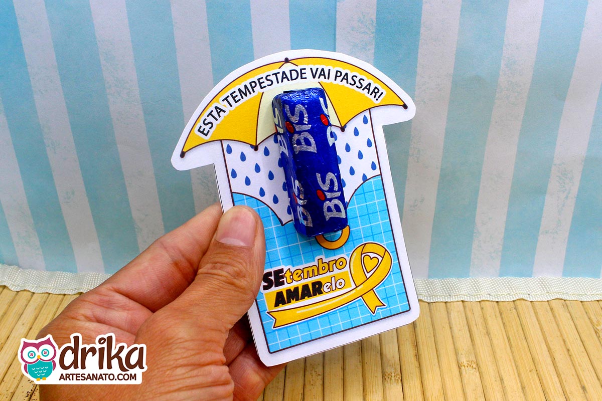 Mão segurando cartão de papel com estampa de grade azul e guarda-chuva amarelo, com um chocolate Bis fixado