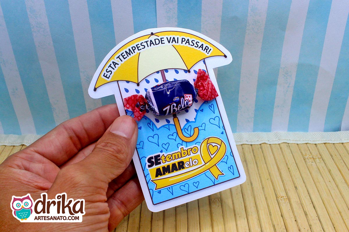 Mão segurando cartão de papel com estampa de corações azuis e guarda-chuva amarelo, com uma bala 7Belo fixada